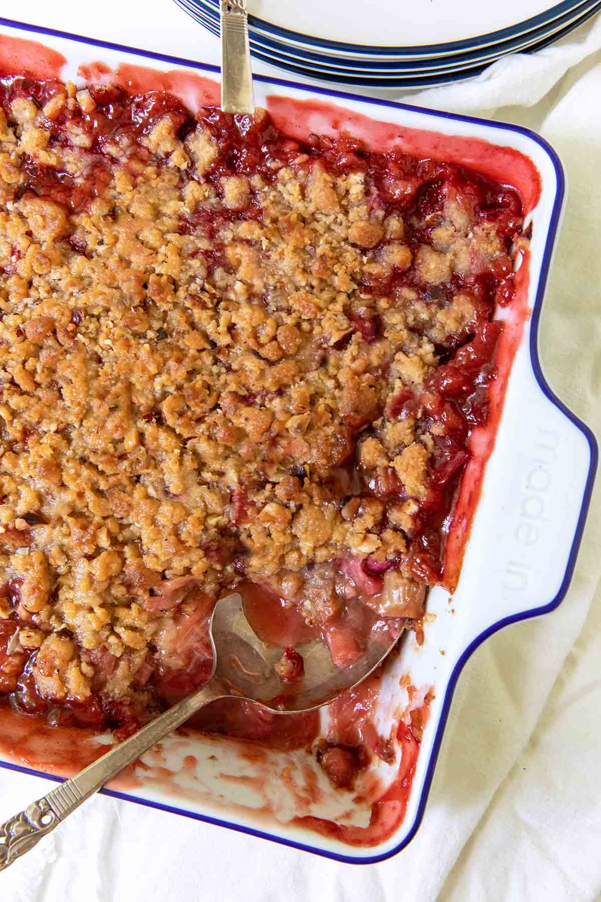 Eine quadratische Auflaufform, gefüllt mit Erdbeer-Rhabarber-Crumble mit Balsamico-Beträufeln, mit einer Portion auf einem Teller und ein paar Erdbeeren als Beilage.