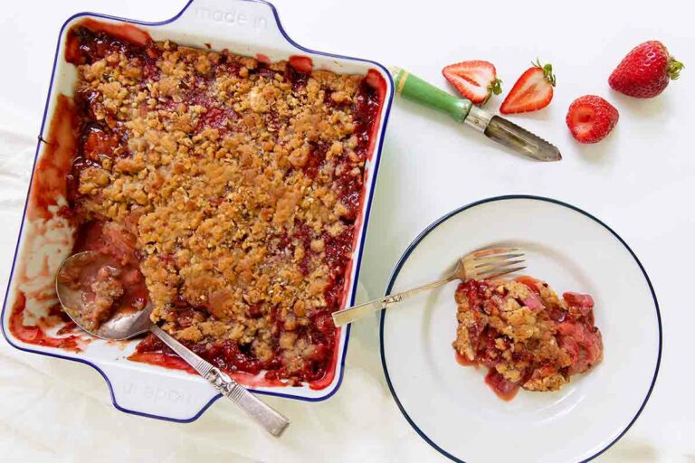 A square baking dish filled with strawberry rhubarb crumble with balsamic drizzle, with a portion on a plate and a few strawberries on the side.