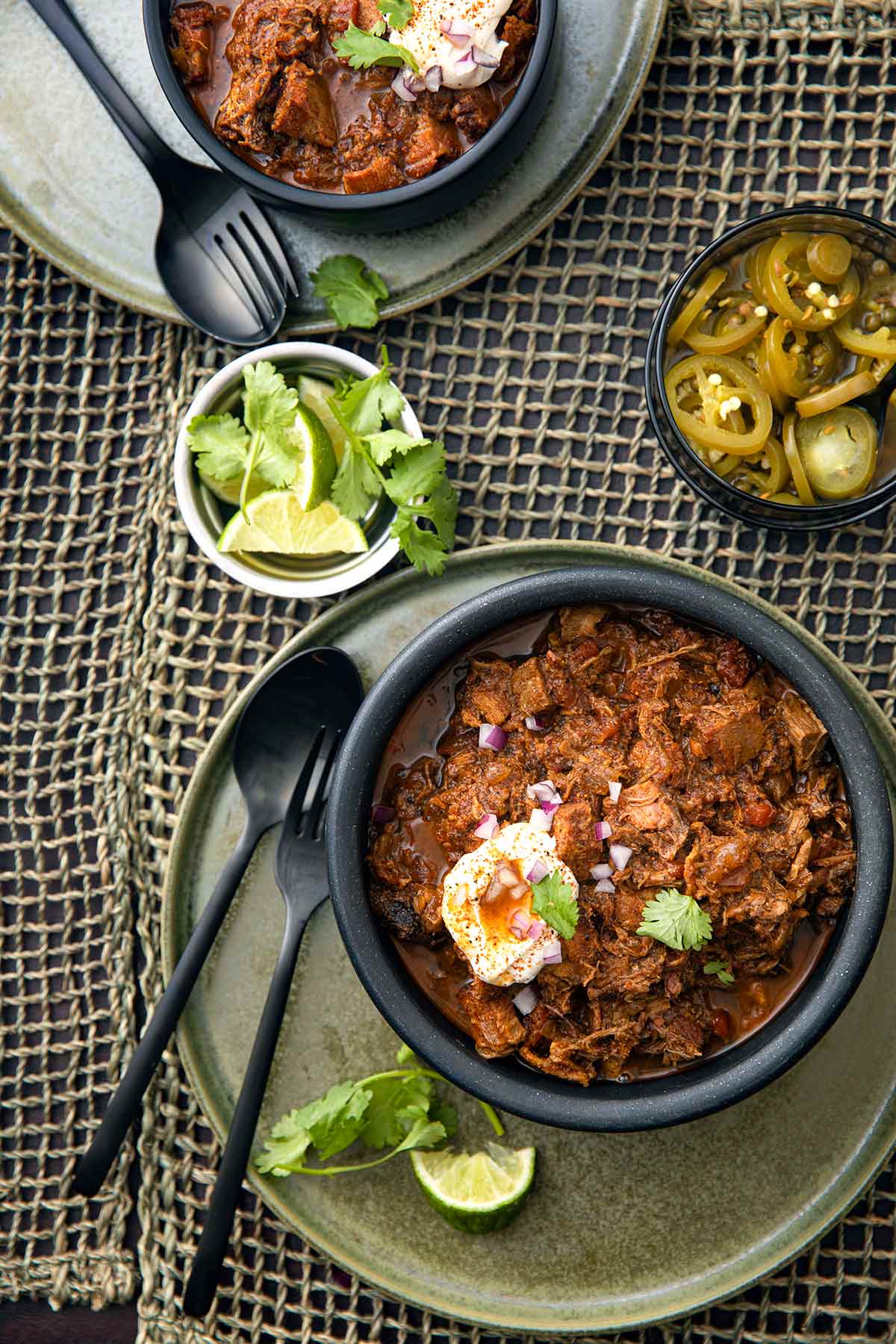 En skål med chili i Texas-stil med fläsk och bringa med en gaffel, sked och skålar med lime, koriander och inlagd jalapenos på sidan.