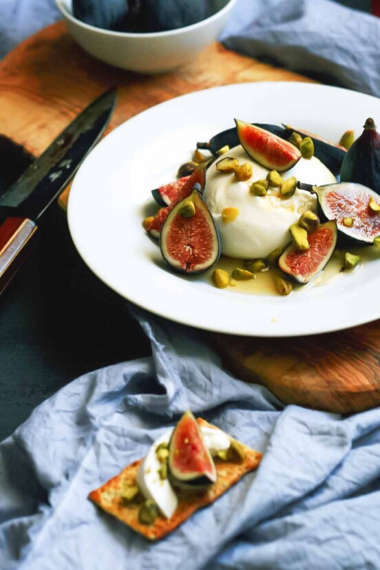 A white plate topped with torn figs and burrata cheese and chopped pistachios with a knife on the side.