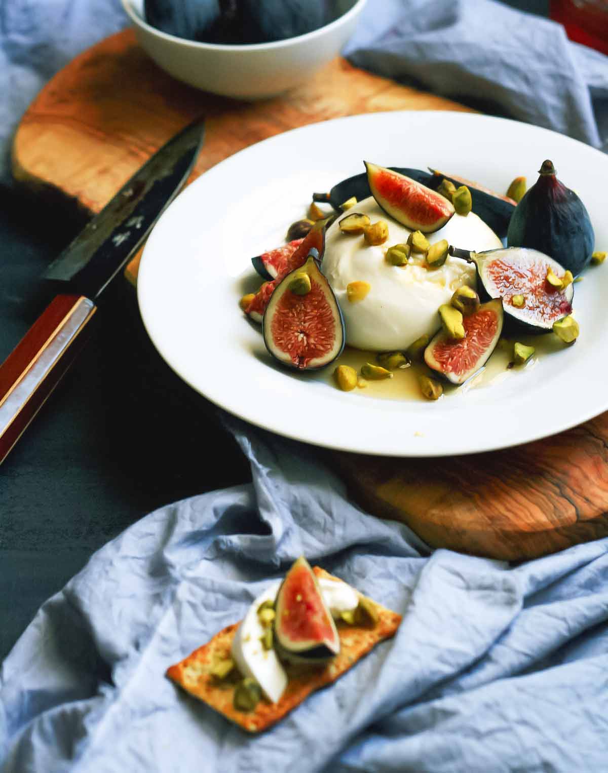 A white plate topped with torn figs and burrata cheese and chopped pistachios with a knife on the side.