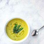 A white bowl filled with vegan asparagus soup with white beans with asparagus tips, ground pepper, and a drizzle of oil on top.