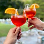 2 hands, each holding a wine glass 3 quarters full of a bubbly orange cocktail with ice and an orange slice.