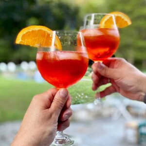 2 hands, each holding a wine glass 3 quarters full of a bubbly orange cocktail with ice and an orange slice.