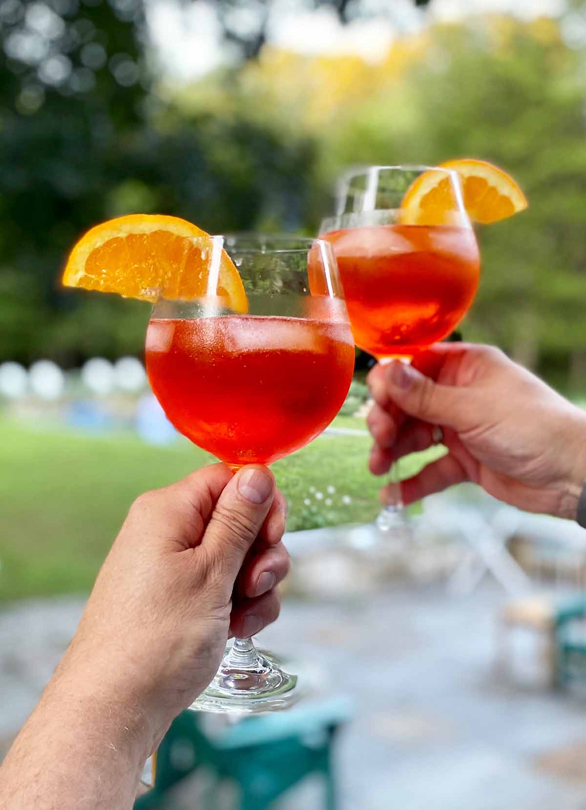 2 hands, each holding a wine glass 3 quarters full of a bubbly orange cocktail with ice and an orange slice.