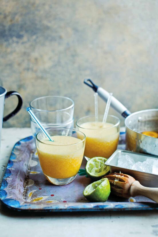 A metal serving tray with two apricot daiquiris with stir sticks, a juiced lemon, and a pot with poached apricots.