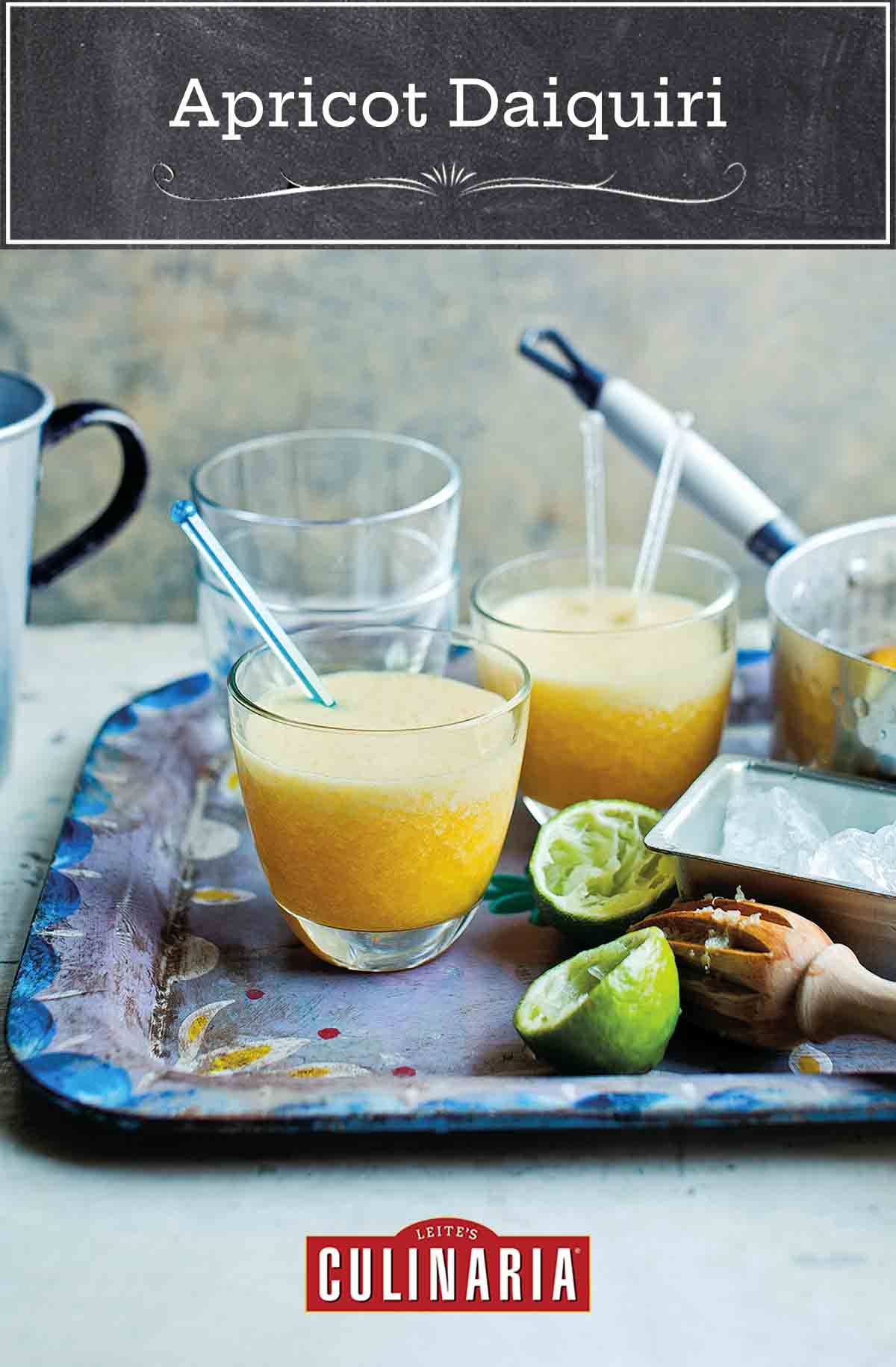 A metal serving tray with two apricot daiquiris with stir sticks, a juiced lemon, and a pot with poached apricots.