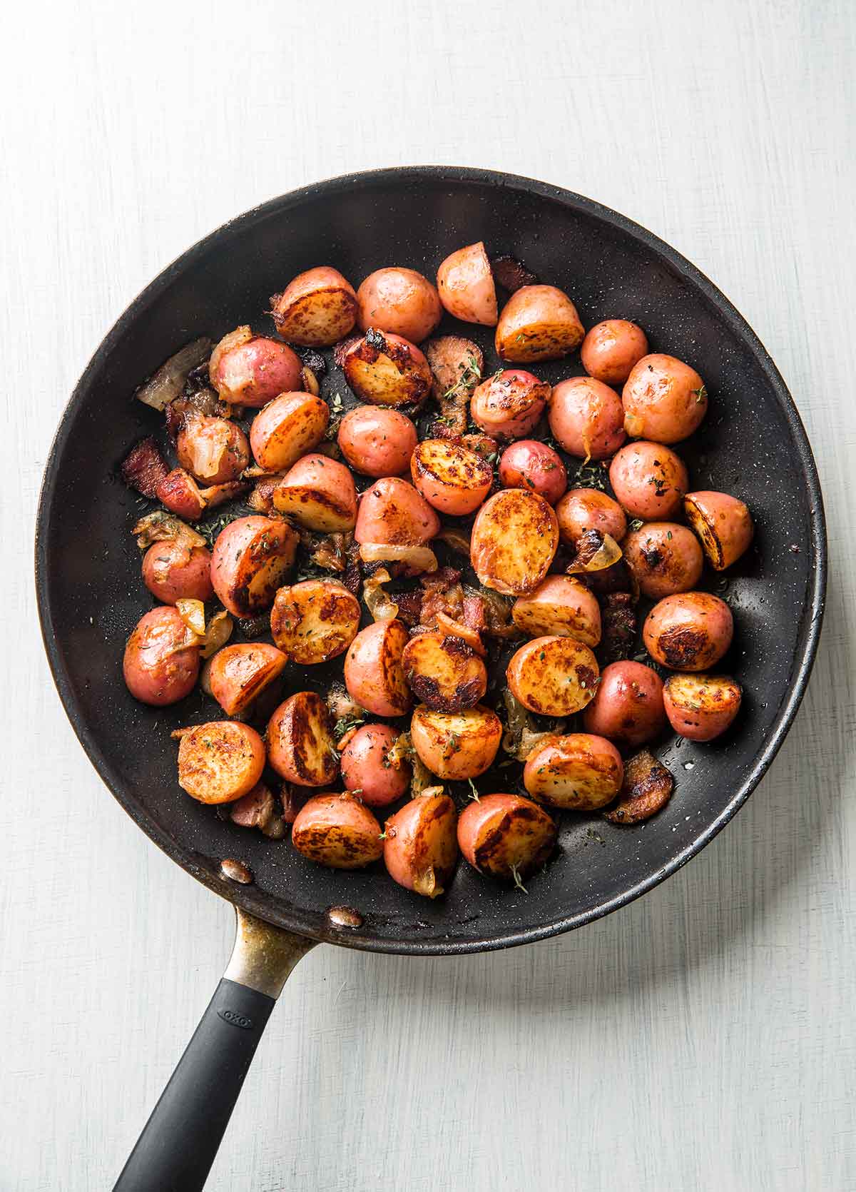 A frying pan filled with halved red potatoes that are browned and covered with crispy bacon and thyme.