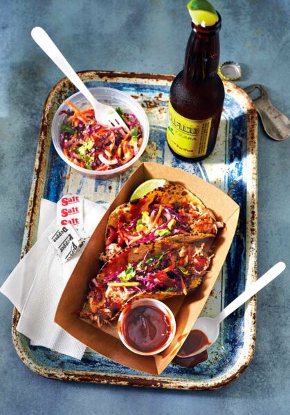 Two BBQ pulled pork tacos in a cardboard dish with a bear and a dish of coleslaw on the side.