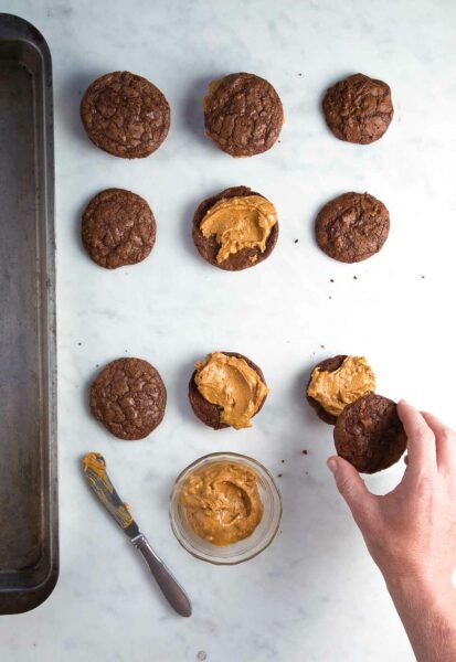 Nine brownie cookies, half without tops and just peanut butter filling. A bowl of filling, a knife and a hand placing a cookie top are also in the picture.