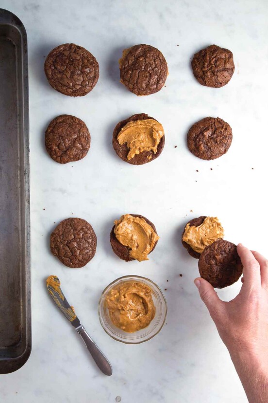 Nine brownie cookies, half without tops and just peanut butter filling. A bowl of filling, a knife and a hand placing a cookie top are also in the picture.