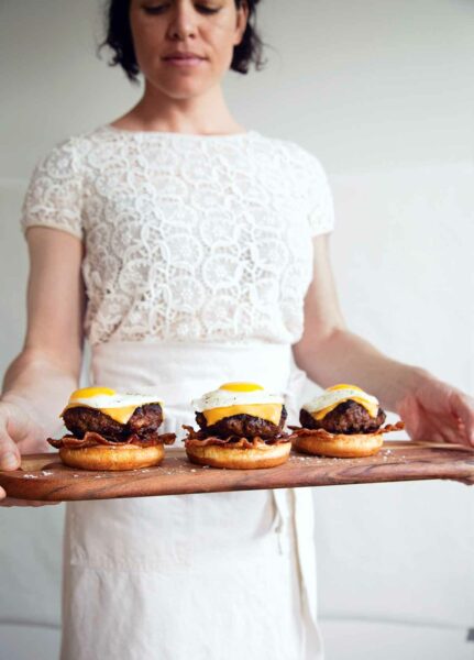 A woman holding a wooden tray with 3 breakfast burgers topped with eggs.