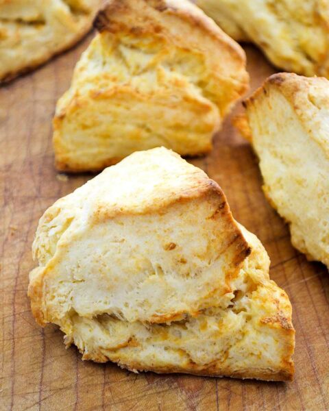Five irregular shaped cheddar biscuits with golden edges, on a wooden cutting board.