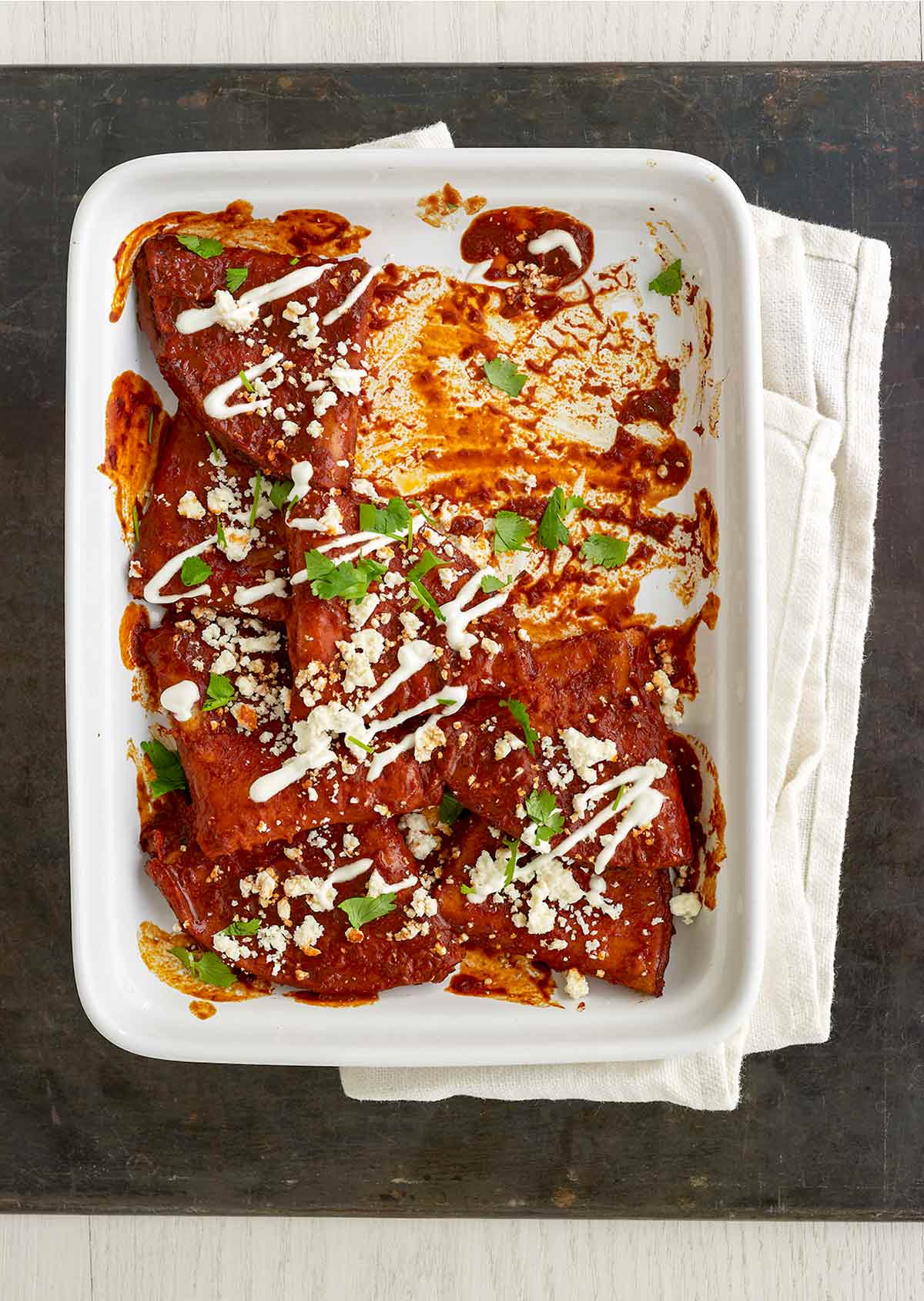 A white casserole dish partially filled with cheese enchiladas, garnished with cilantro, cheese, and crema.