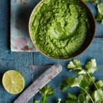 A bowl of bright green cilantro chutney on a blood wooden table with a flower napkin, cilantro leaves, half a lime, and a knife.