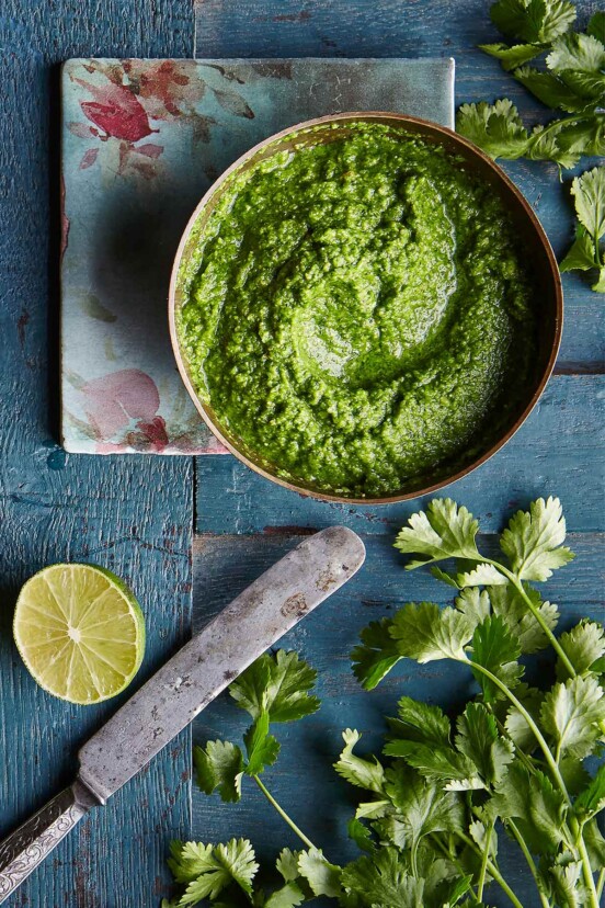 A bowl of bright green cilantro chutney on a blood wooden table with a flower napkin, cilantro leaves, half a lime, and a knife.