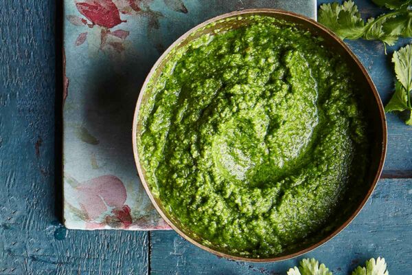 A bowl of bright green cilantro chutney on a blood wooden table with a flower napkin and cilantro leaves.