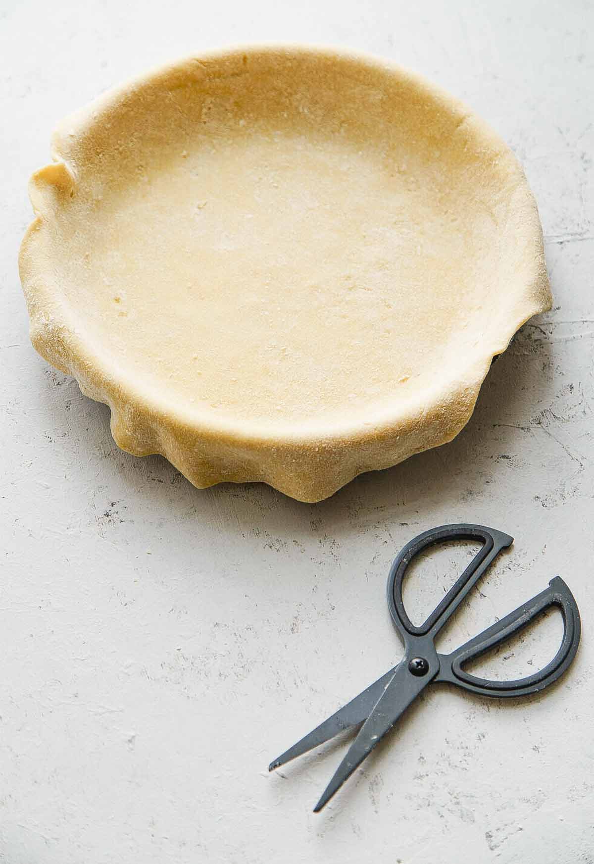 Empty pie crust in a pie plate, with kitchen shears next to it.