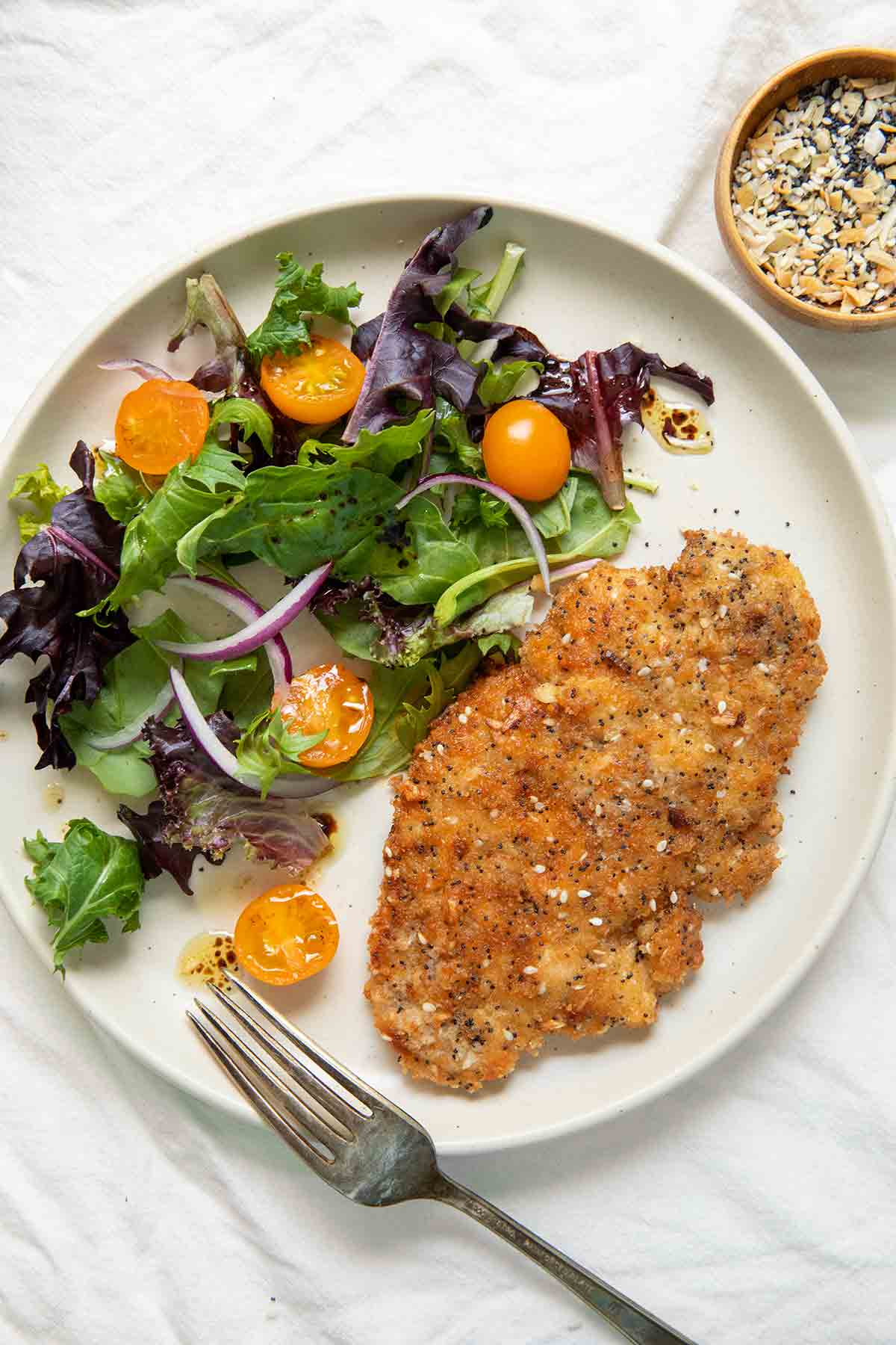 A white plate with a lettuce, cherry tomato, and onion salad, beside a chicken breast coated in crisp breading. A knife and fork lay on the edge of the plate.