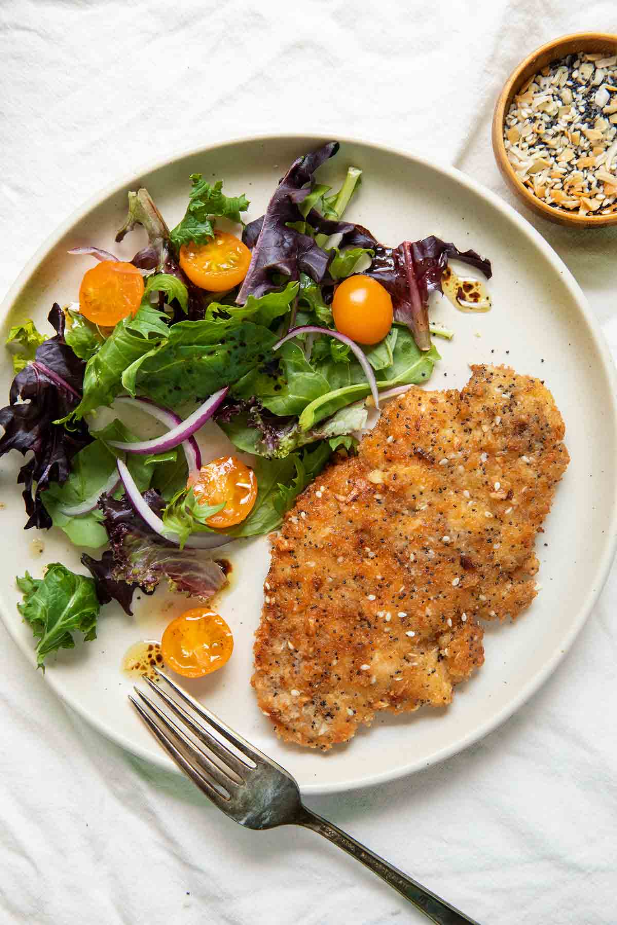 En vit tallrik med sallad, körsbärstomat och löksallad, bredvid ett kycklingbröst täckt med krispig panering.  En kniv och gaffel låg på kanten av tallriken.