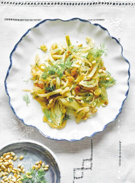 A white and blue bowl filled with fennel, dill, pine nuts, and golden raisins on a white tablecloth.