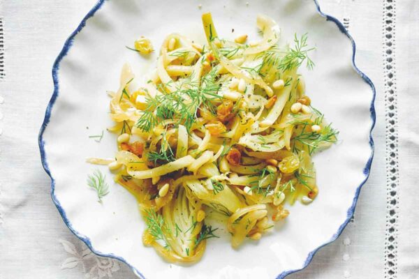 A white and blue bowl filled with fennel, dill, pine nuts, and golden raisins on a white tablecloth.