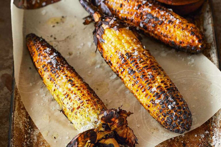 Three cobs of grilled corn with husks attached, lying on a metal sheet pan with parchment paper.Three cobs of grilled corn with husks attached, lying on a metal sheet pan with parchment paper.