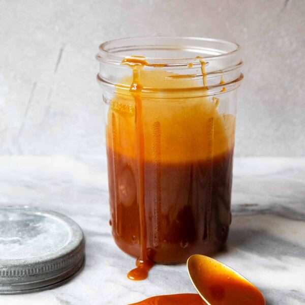 A jar of caramel sauce with some dripping down the side and a spoon in front of the jar.