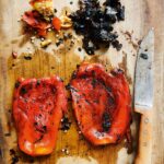 2 roasted red peppers on a cutting board with a large knife, and scraps of charred pepper skin.