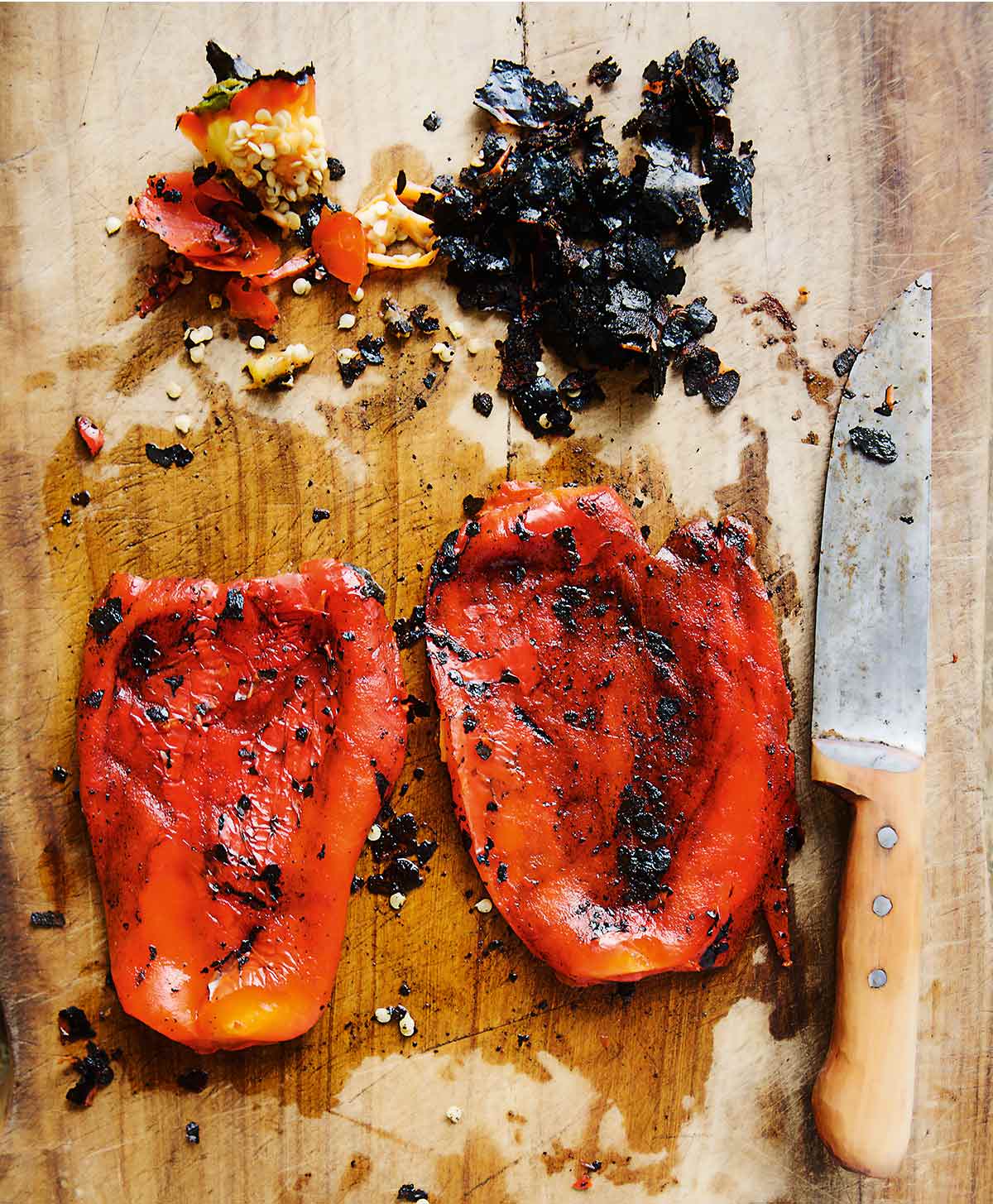 2 roasted red peppers on a cutting board with a large knife, and scraps of charred pepper skin.