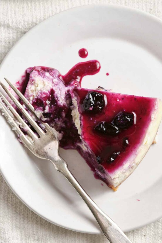 A white plate on a white tablecloth, with a slice of cheesecake topped with blueberry sauce and flanked by a fork.