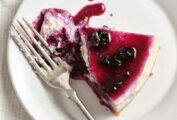A white plate on a white tablecloth, with a slice of cheesecake topped with blueberry sauce and flanked by a fork.
