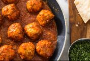 A pan full of turkey meatballs with tomato sauce, beside a chopping board with Parmesan and a bowl of parsley.