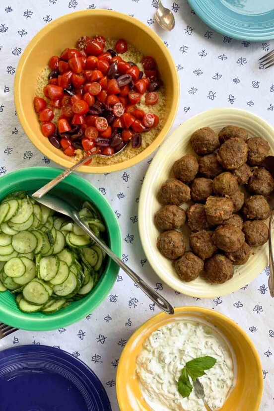 A platter of lamb meatballs with yogurt sauce, tomatoes, and cucumbers in bowls on the side.