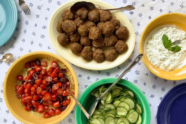 A tabletop with bowls of tomatoes with couscous, sliced cucumber, yogurt sauce, and lamb meatballs.