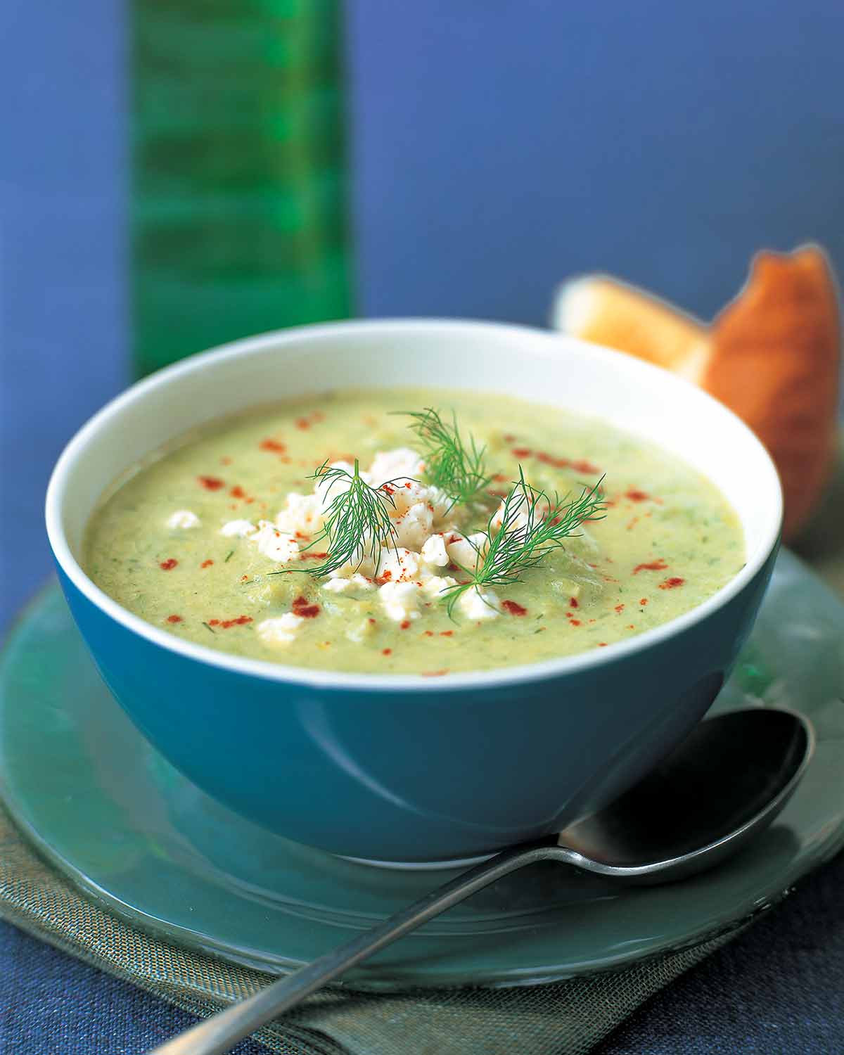 A blue bowl filled with a pale green cream soup, topped with paprika, feta, and fresh dill fronds.