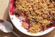 A white bowl filled with blueberry nectarine crisp with a scoop missing and a spoon laying beside it.