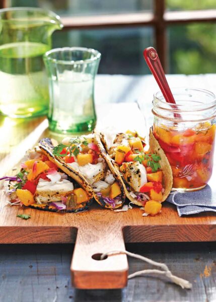 A large wooden cutting board with 3 tacos on it, each filled with grilled fish and peach salsa. A jar of salsa and a spoon are next to them.