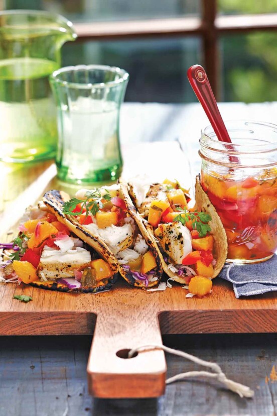 A large wooden cutting board with 3 tacos on it, each filled with grilled fish and peach salsa. A jar of salsa and a spoon are next to them.