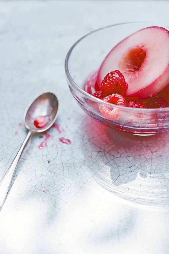 A glass bowl filled with a poached nectarine half and raspberries with a spoon on the side.