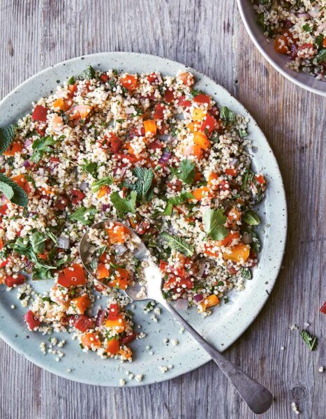 A light blue plate and spoon on a wooden table, filled with quinoa, diced peppers, tomato, parsley, mint, and red onion.