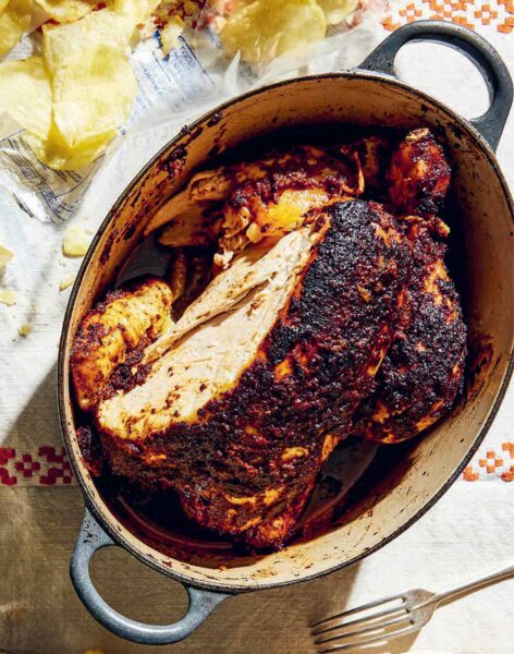 A cast-iron Dutch oven filled with a deeply roasted chicken, flanked by a bag of potato chips.