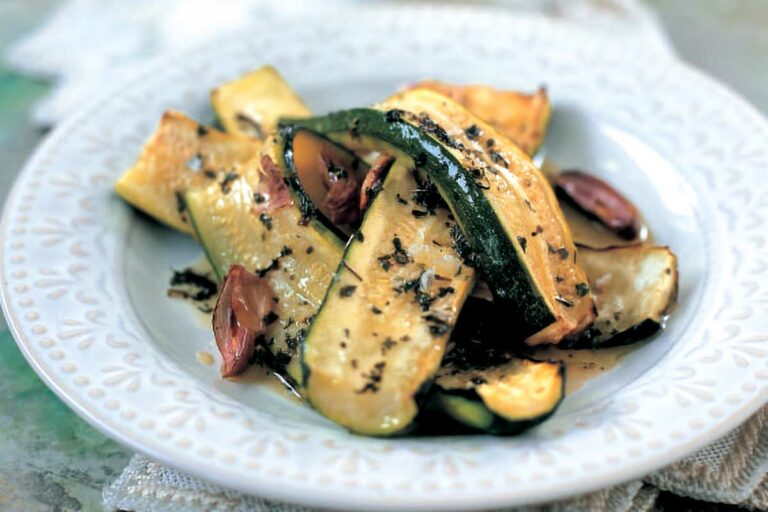 A white plate filled with slices of roasted zucchini, olive oil, dark roasted cloves of garlic, and mint.