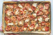 A sheet pan covered with parchment paper, filled with layers of sliced zucchini, tomatoes, shredded mozzarella, piles of ricotta and marinara sauce.