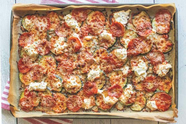 A sheet pan covered with parchment paper, filled with layers of sliced zucchini, tomatoes, shredded mozzarella, piles of ricotta and marinara sauce.