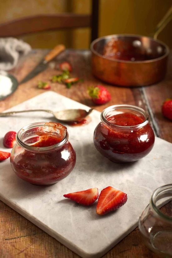 Two glass jars filled with deeply red strawberry jam surrounded by fresh berry slices.