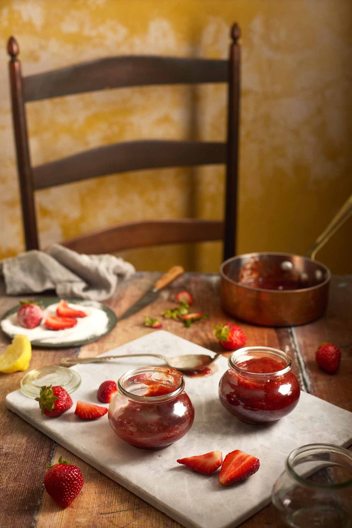 Two jars of small batch strawberry jam on a marble board with fresh sliced strawberries and a copper pot.