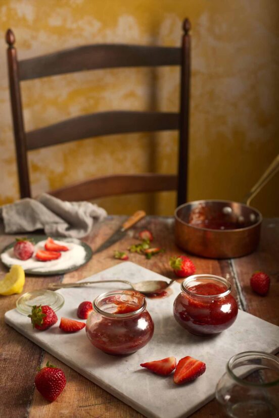 Two jars of small batch strawberry jam on a marble board with fresh sliced strawberries and a copper pot.