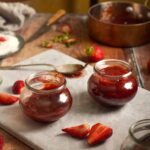 Close-up of two glass jars filled with ruby red strawberry jam surrounded by fresh berry slices.