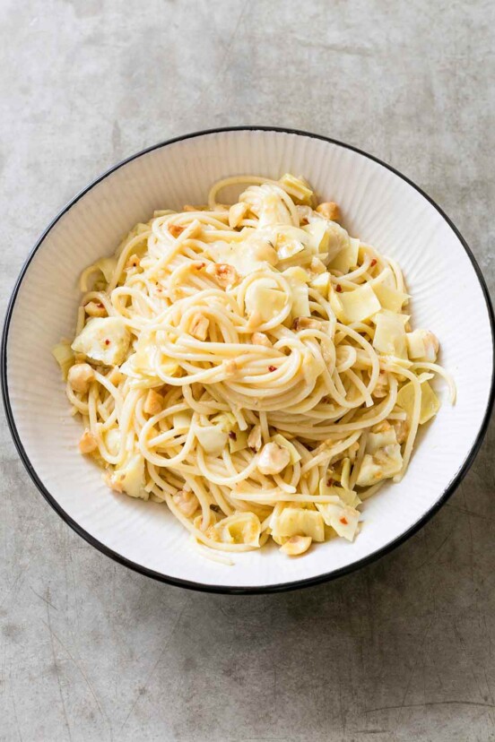 A big white bowl filled with spaghetti, chunks of artichokes, hazelnuts, flecks of chile pepper flakes, and Parmesan cheese.